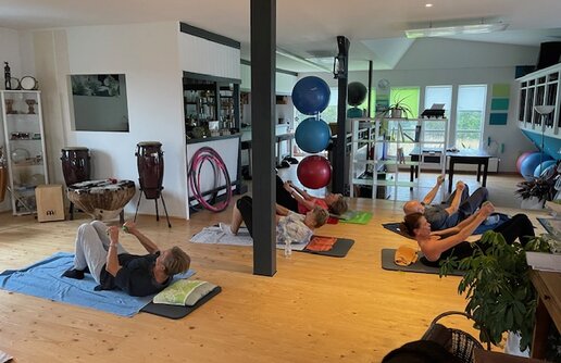 Funktionstraining: In einem hellen Raum mit Holzboden führen mehrere Personen eine Gymnastikübung auf Matten am Boden aus.