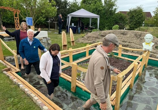 vier Personen die in das Wasserbecken Barfuß einsteigen, im Hintergrund steht ein Pavillon und eine Büste
