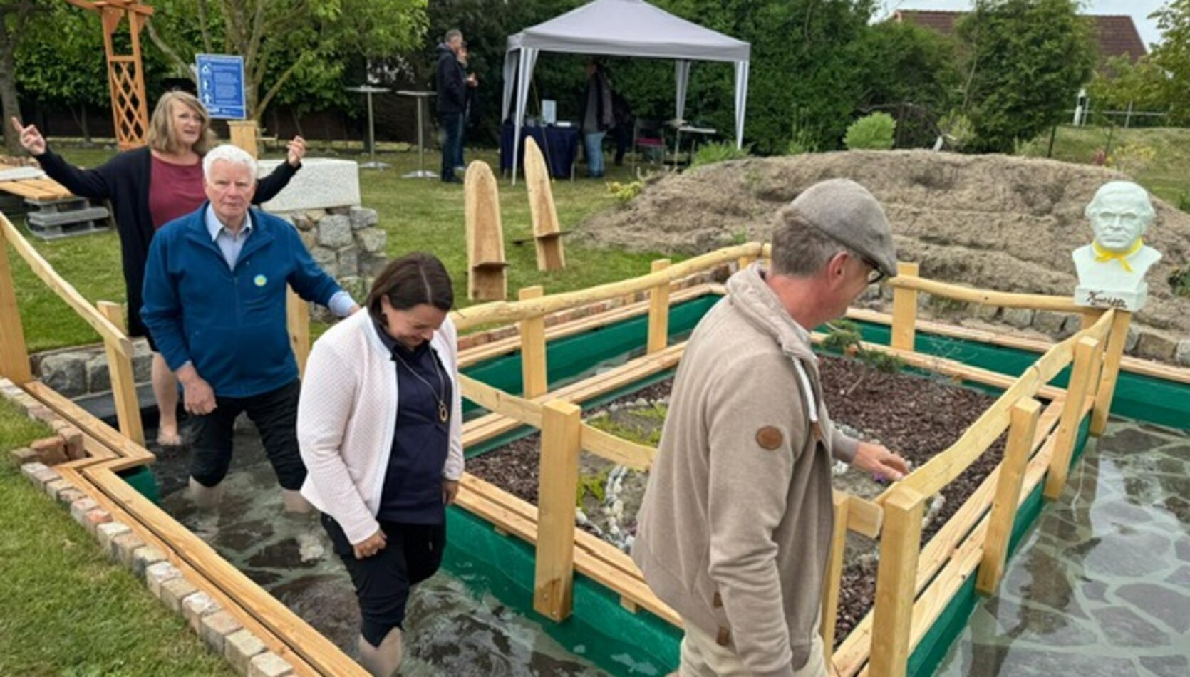 vier Personen die in das Wasserbecken Barfuß einsteigen, im Hintergrund steht ein Pavillon und eine Büste